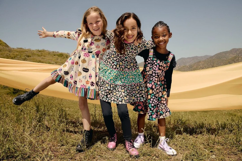 Three cheerful girls in floral dresses pose outdoors in a grassy field on a sunny day, wearing colorful patterned outfits with mountains visible.