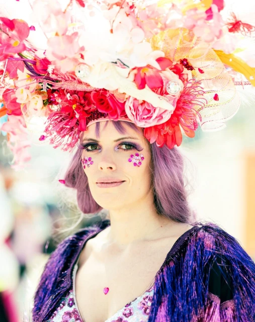 Woman with purple hair and flower crown, wearing fringed sequin top, looks at the camera with a slight smile, against soft bokeh.