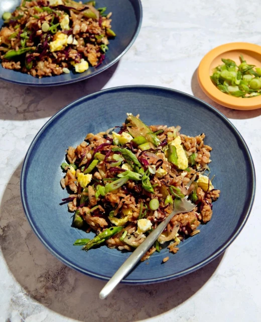 Close-up of two blue bowls filled with fried rice, vegetables, and eggs, with a fork in one bowl, presented on a marble surface.