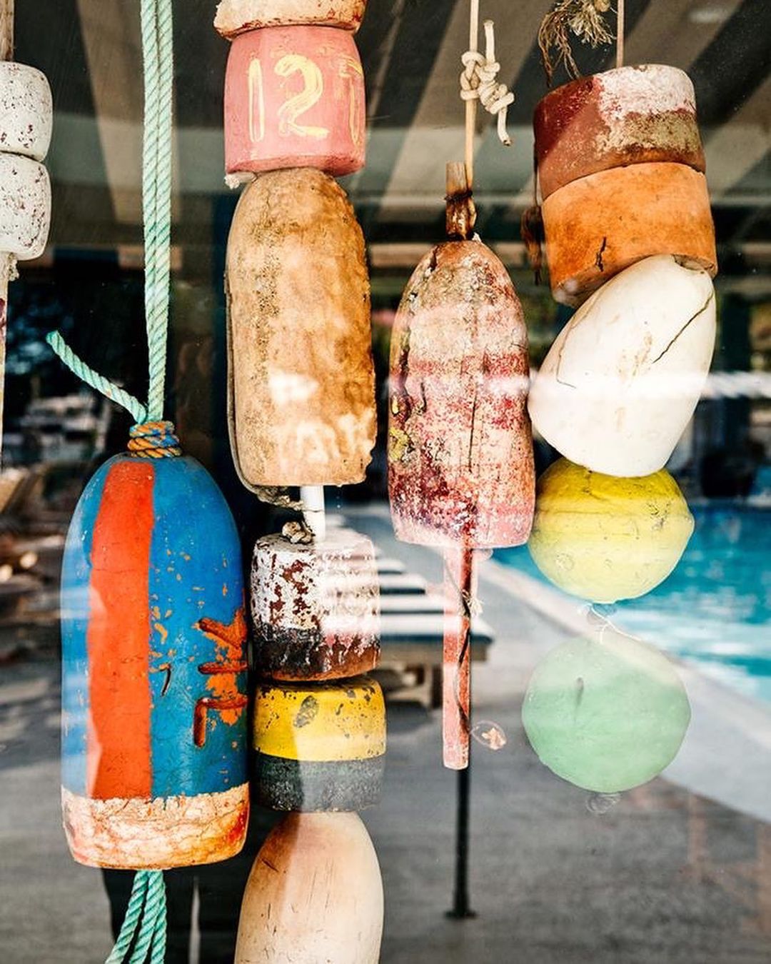 Colorful lobster buoys hang in a window display with a swimming pool visible through the glass, creating a vibrant and nautical scene.
