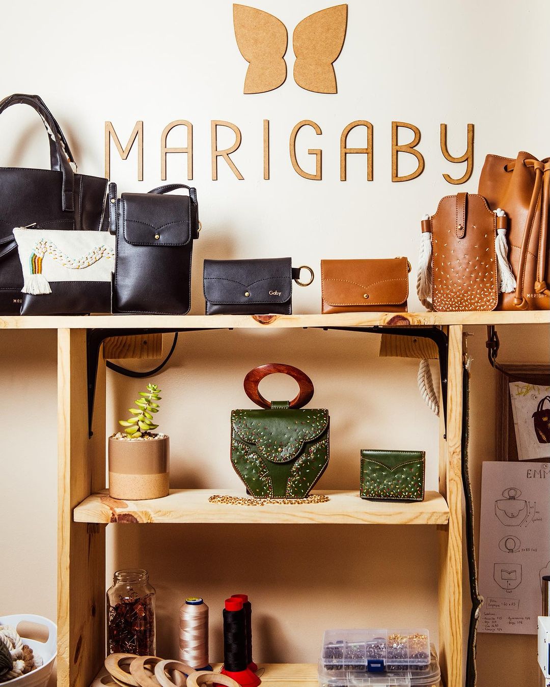 Display of handcrafted leather handbags in neutral colors on a wooden shelf with a butterfly logo in the background.