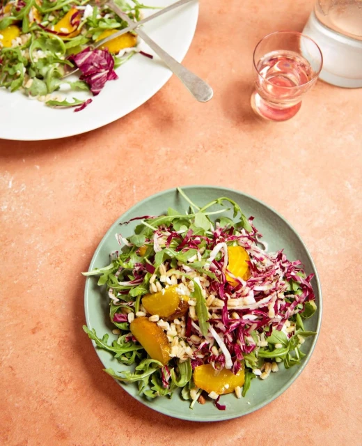 Overhead shot: fresh salad on green plate with arugula, radicchio, orange segments and grains, alongside drink and plate on coral surface.