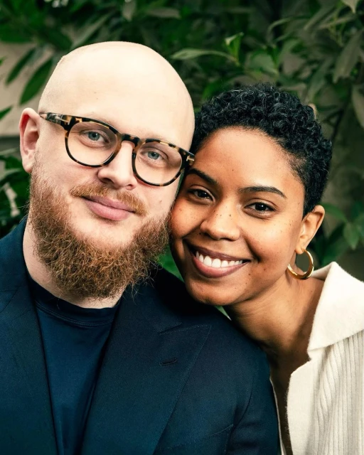 Portrait of a stylish couple, a man with glasses and a beard next to a woman with short hair, both smiling, in front of green leaves.