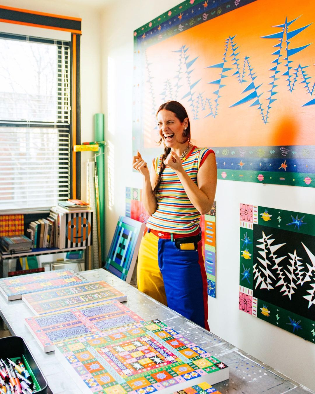 A woman laughs in a colorful art studio, surrounded by paintings and art supplies, with bright natural light streaming through the window.