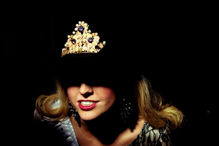 Woman with blonde hair wearing a jeweled gold crown; upper body lit against a black background. Beauty and fashion content.