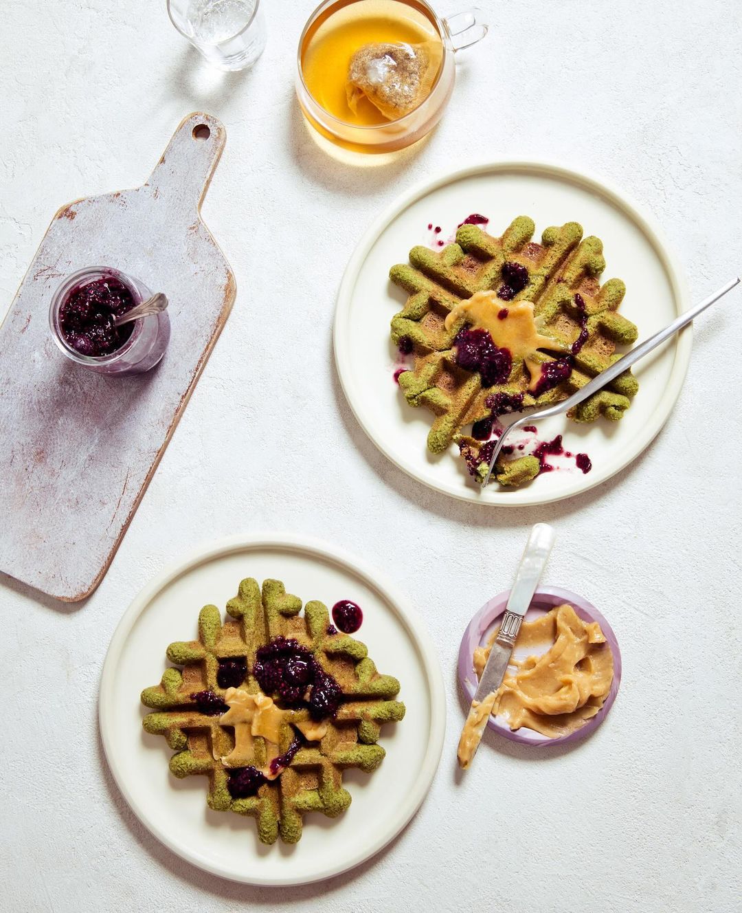 Overhead shot of two green waffles on plates topped with peanut butter and berry jam, tea, and a cutting board with jam.