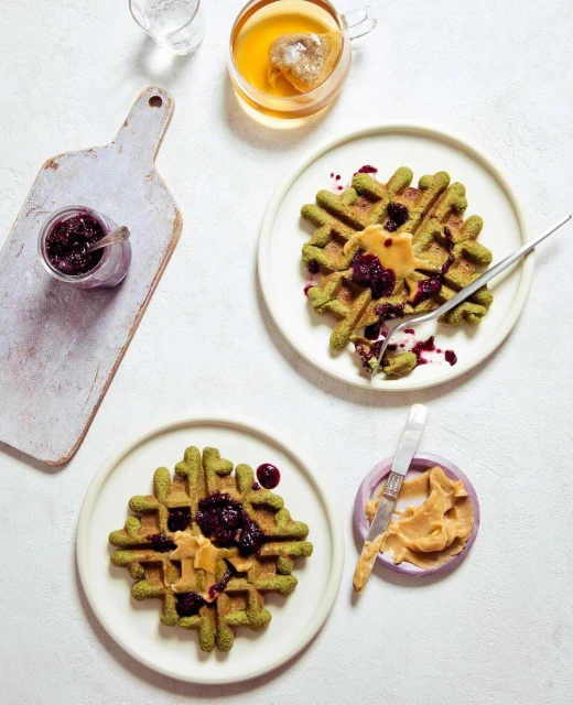 Overhead shot of two green waffles on plates topped with peanut butter and berry jam, tea, and a cutting board with jam.