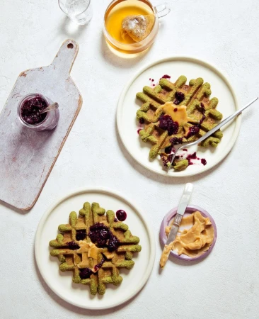 Overhead shot of two green waffles on plates topped with peanut butter and berry jam, tea, and a cutting board with jam.