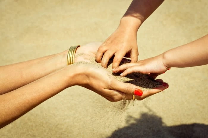 Close-up view of an adult and child's hands playing with sand on a tan-colored beach outdoors during daylight, creating a heartwarming scene.