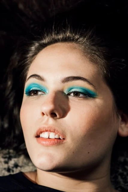 Close-up portrait of a young adult woman with bright turquoise eyeshadow, fair skin, and dark hair, against a dark background.