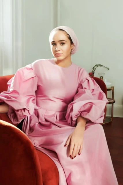 Young woman in a pink gown and headscarf sits in a red chair with a neutral backdrop. Fashionable attire and graceful pose.