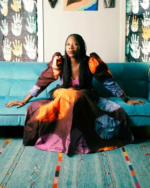 A Black woman with braided hair wearing an orange, purple, and brown dress is seated on a blue sofa in an indoor setting.