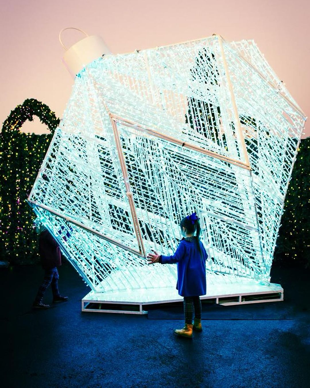 Young girl in blue coat reaching toward large illuminated Christmas ornament structure in outdoor park setting with green holiday foliage.