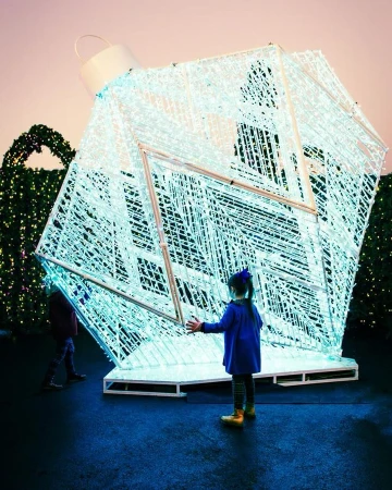 Young girl in blue coat reaching toward large illuminated Christmas ornament structure in outdoor park setting with green holiday foliage.