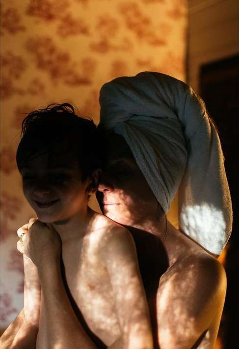 Sunlight filters through window casting dappled light on a woman in towel wrap hugging a child, warm indoor scene, family aesthetic.