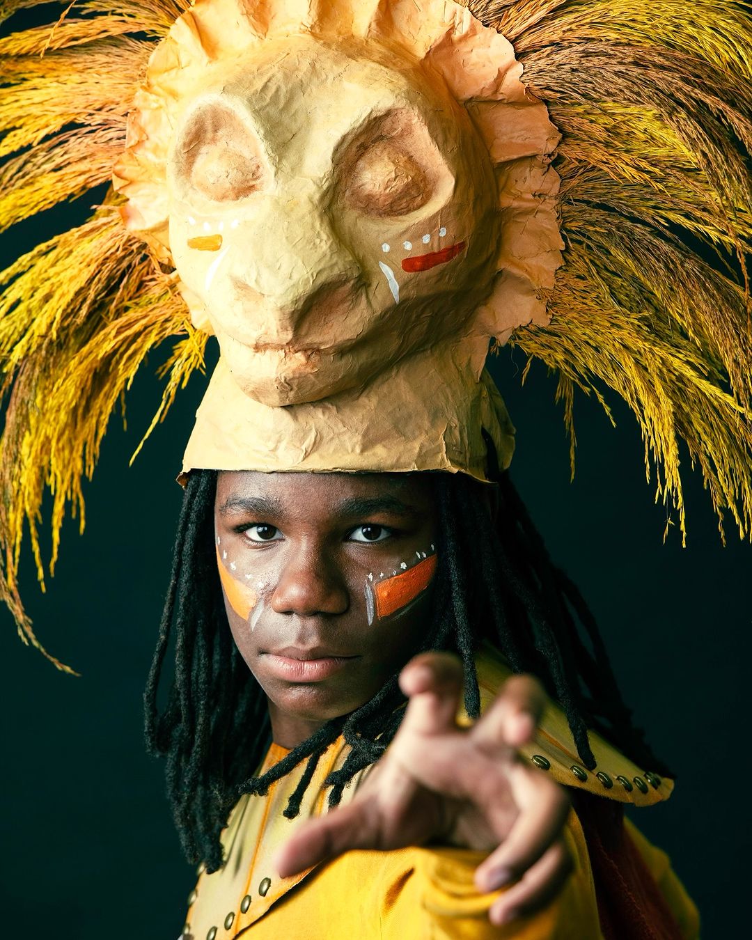 Teen with lion mask headdress and face paint posing against a dark background, artistic portrait with focus on character and detail.