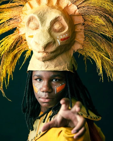Teen with lion mask headdress and face paint posing against a dark background, artistic portrait with focus on character and detail.