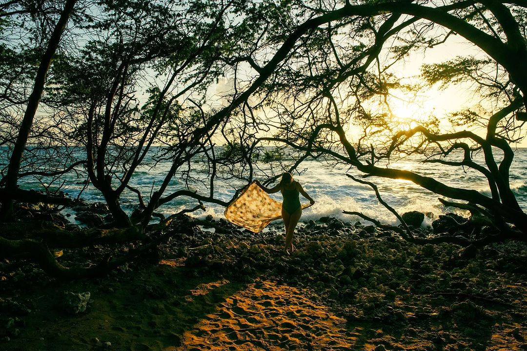 Woman in green swimsuit holding a patterned scarf on a rocky beach framed by silhouetted tree branches at sunset.