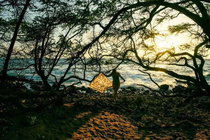 Woman in green swimsuit holding a patterned scarf on a rocky beach framed by silhouetted tree branches at sunset.