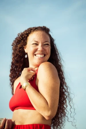 Smiling woman with long curly brown hair and freckles wearing a red top against a clear blue sky background.