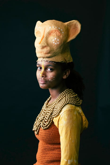 Young woman in a headshot wearing a textured yellow top, beaded necklace, and handcrafted feline mask in front of a dark background.