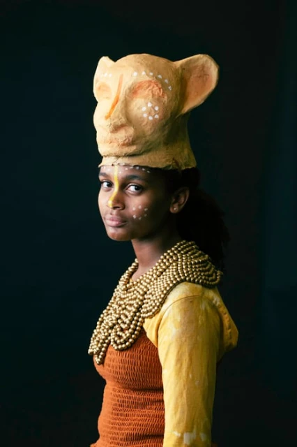 Young woman in a headshot wearing a textured yellow top, beaded necklace, and handcrafted feline mask in front of a dark background.