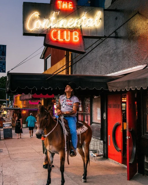 Adult man on mule in urban scene. Neon sign reads 'The Continental Club'. The man wears a t-shirt, blue jeans, and a black hat.