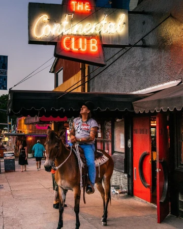 Adult man on mule in urban scene. Neon sign reads 'The Continental Club'. The man wears a t-shirt, blue jeans, and a black hat.