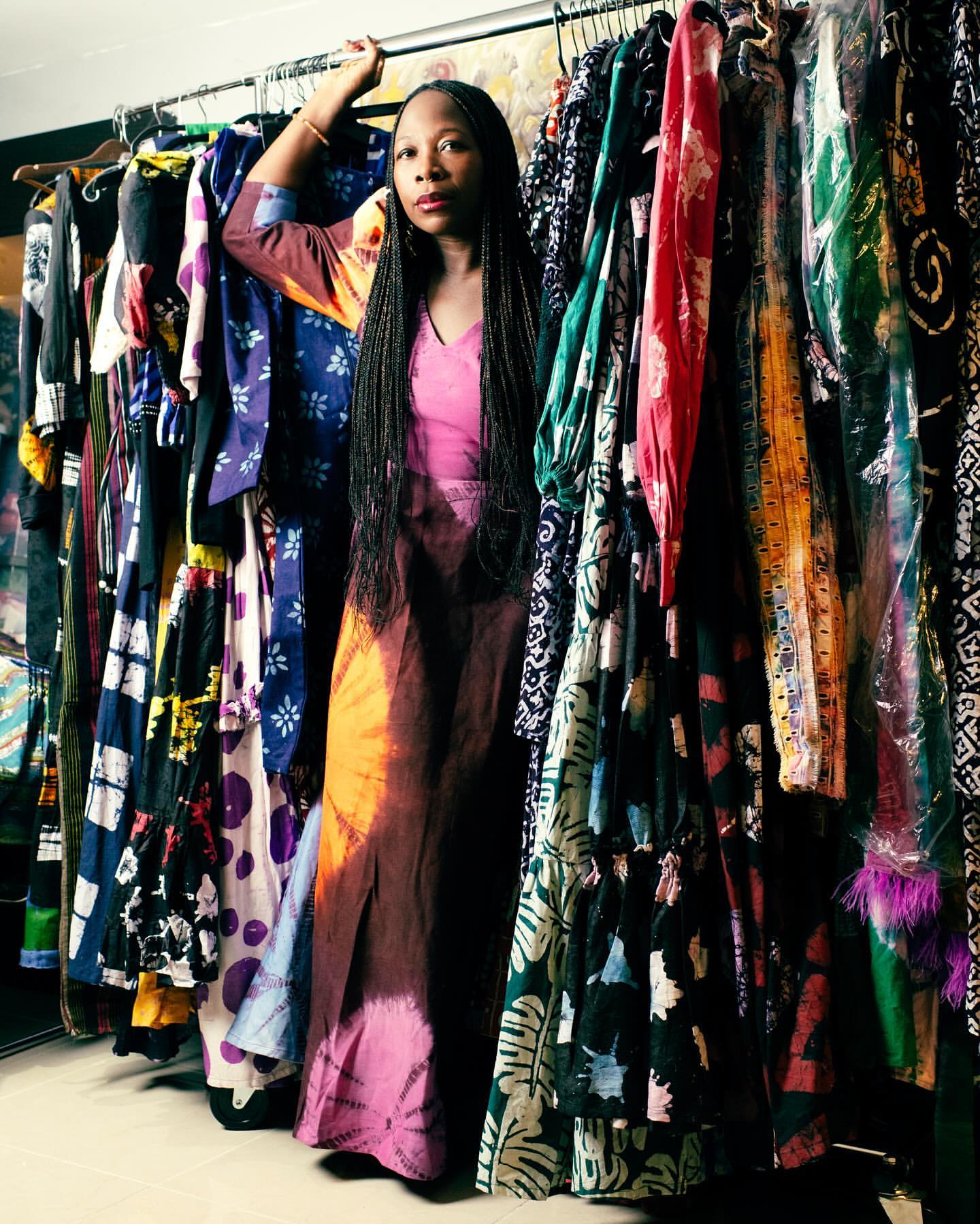 Fashion photo of a woman standing in front of a rack of dresses, posing in a colorful tie-dye gown in an indoor setting with muted lighting.