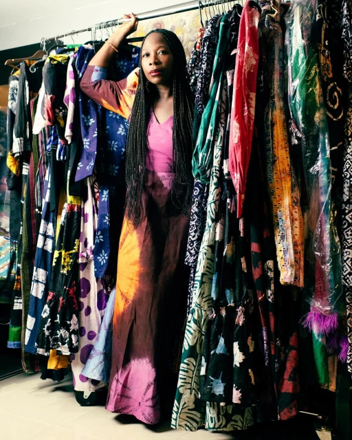 Fashion photo of a woman standing in front of a rack of dresses, posing in a colorful tie-dye gown in an indoor setting with muted lighting.