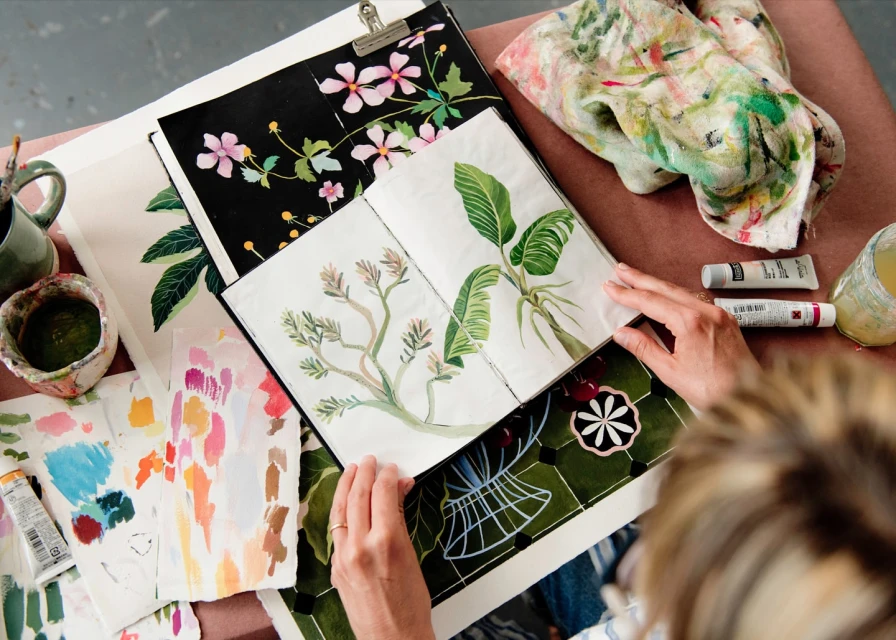 Artist working with sketchbooks featuring floral and botanical illustrations, with watercolors and brushes in a light-filled studio.