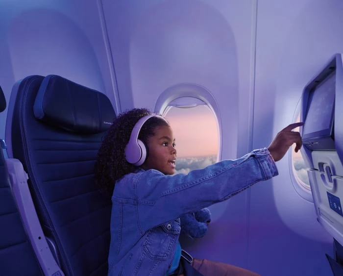 A young girl with headphones in an airplane seat enjoys the entertainment system by the window, bathed in cool, violet lighting.