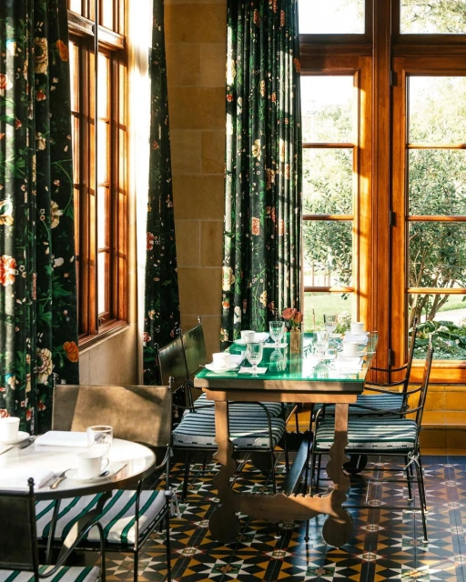 A dining room with a green glass-topped table set for a meal, surrounded by chairs, windows, and floral curtains, showcasing an elegant interior.