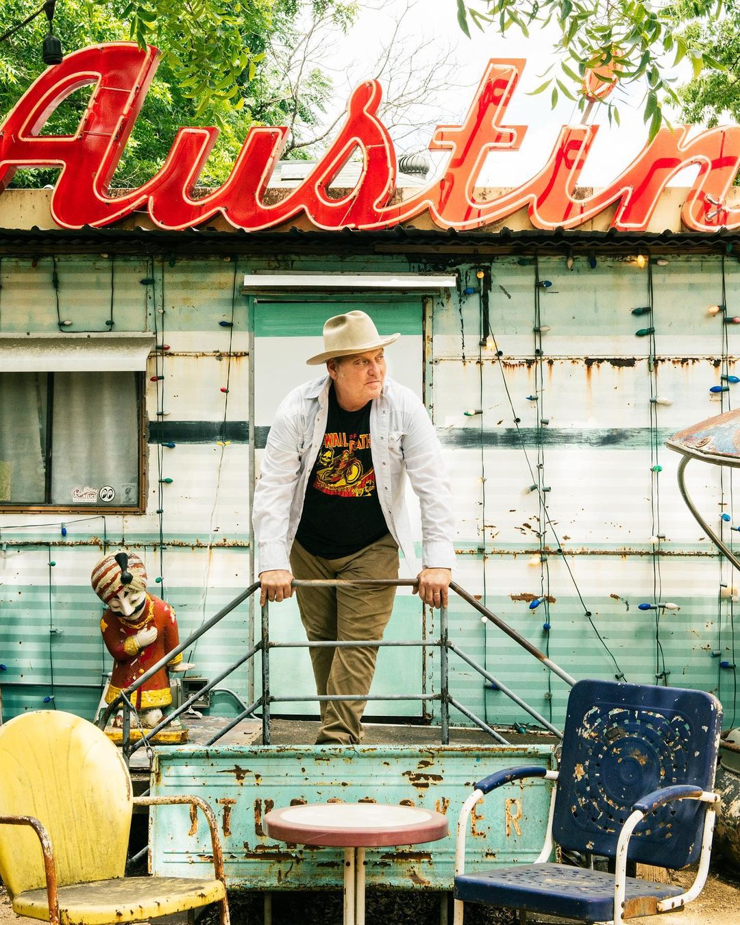 Man in cowboy hat stands on vintage trailer in front of 'Austin' sign, surrounded by old chairs, captured in medium shot outdoors.