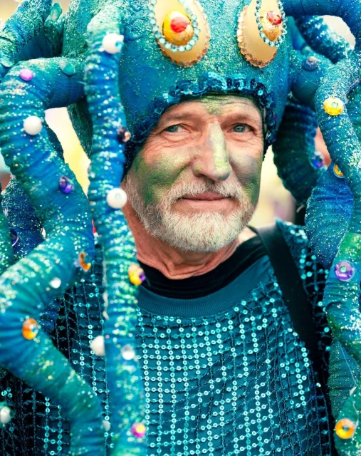 Close-up of an adult male in a teal sequin top, green face paint, and an octopus hat with blue tentacles and colorful beads.