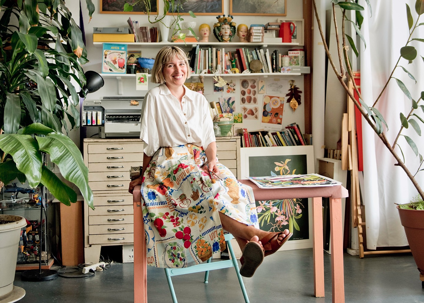 Smiling woman with blonde hair sits on a turquoise chair in a sunlit artist studio, wearing a white shirt and colorful skirt.