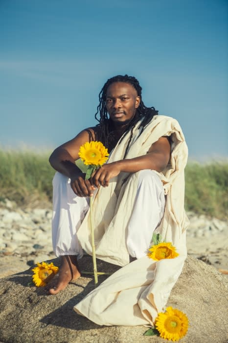 An adult African-American man sits on a sandy mound outdoors, holding sunflowers and wearing light-colored clothing under a clear sky.
