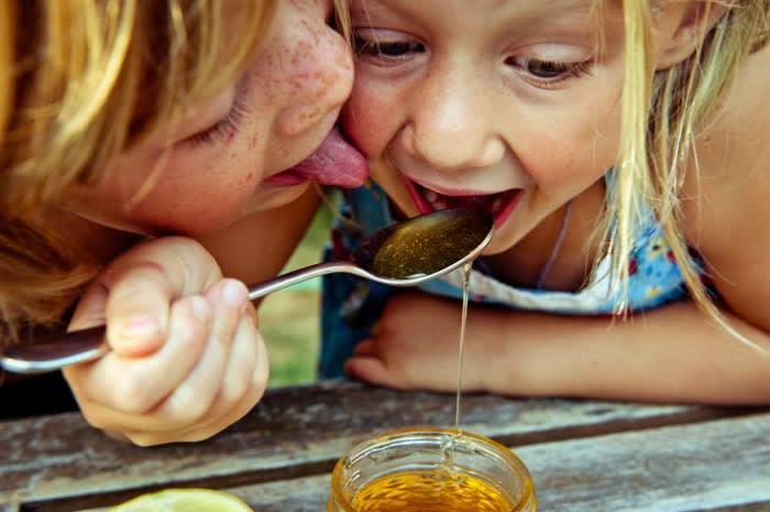 Two children sharing honey from a spoon, one child eating from the spoon while the other licks the spoon. Close-up, happy expression.
