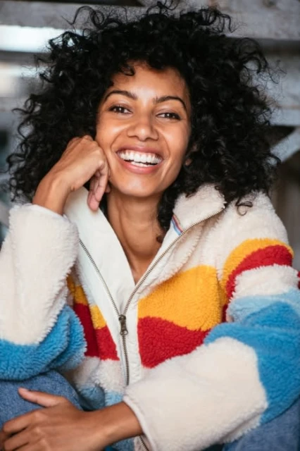 Smiling Black woman in a colorful sherpa jacket sitting indoors, looking at the camera with a happy expression and curly hair.