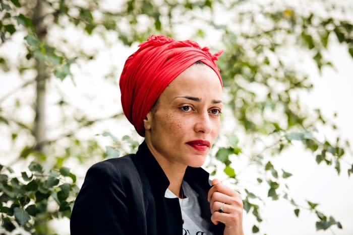 Woman with a red turban and black blazer stands outdoors against a leafy backdrop, looking to the side with a neutral facial expression.