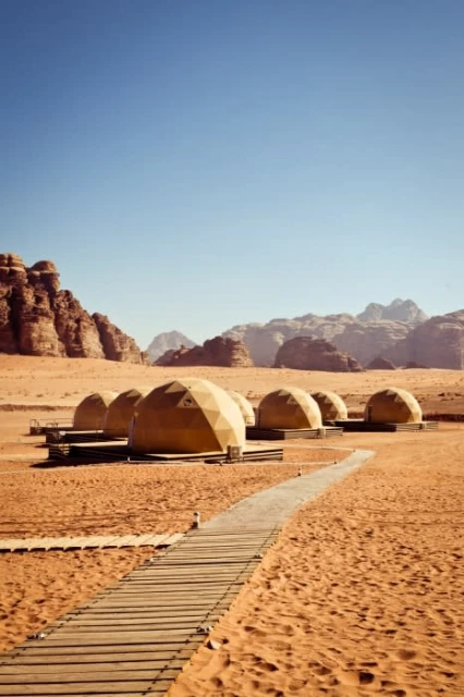 Desert glamping resort with domed tents, wooden walkway, and mountains in the background on a clear day in Wadi Rum, Jordan.