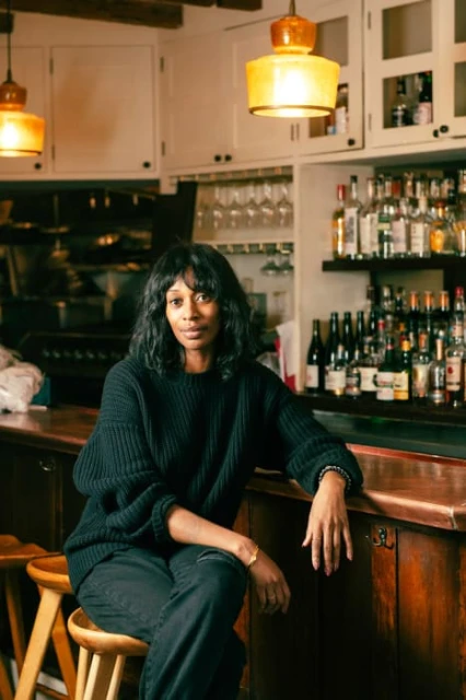 Woman with dark hair wearing a black sweater sitting on a wooden stool in front of a bar with bottles of liquor in the background.