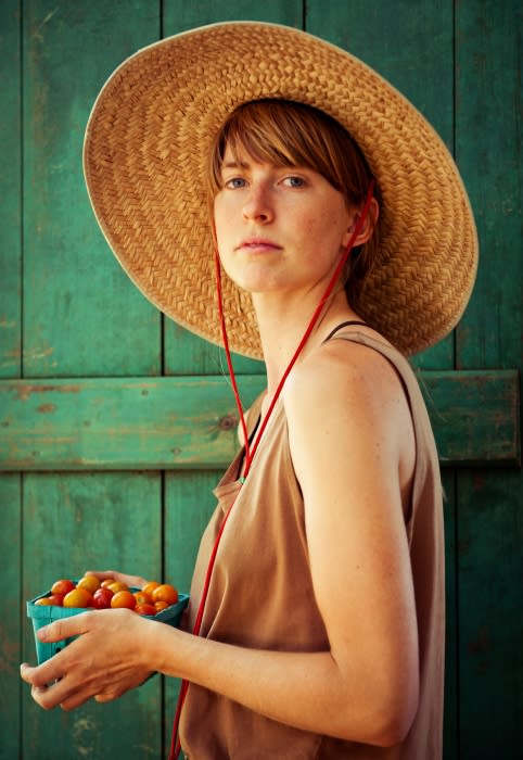 A young woman in a straw hat holds a basket of cherry tomatoes against a weathered green door, captured in a medium shot.