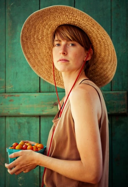 A young woman in a straw hat holds a basket of cherry tomatoes against a weathered green door, captured in a medium shot.