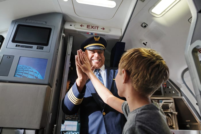 Airplane pilot high-fives a young boy at the entrance to the aircraft; the pilot wears a blue uniform and hat, child wears a gray shirt.