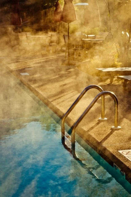 Steam rises from a swimming pool with a pool ladder. Lounge chairs and tables with umbrellas stand on the concrete deck.