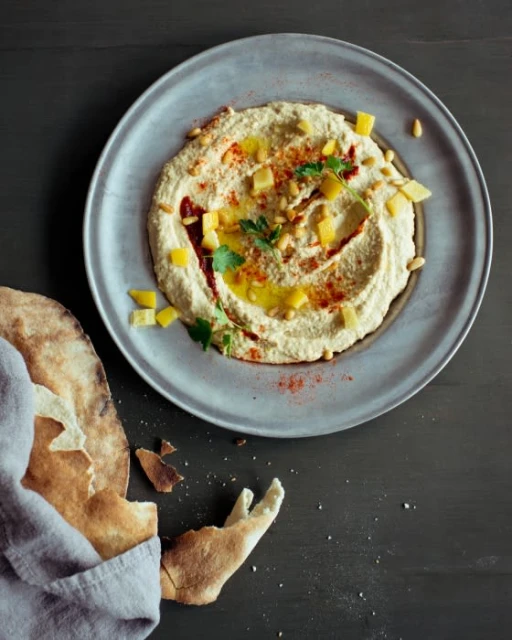 Overhead shot of hummus garnished with mango chunks, paprika, olive oil, and herbs on a gray plate with pita bread and linen.