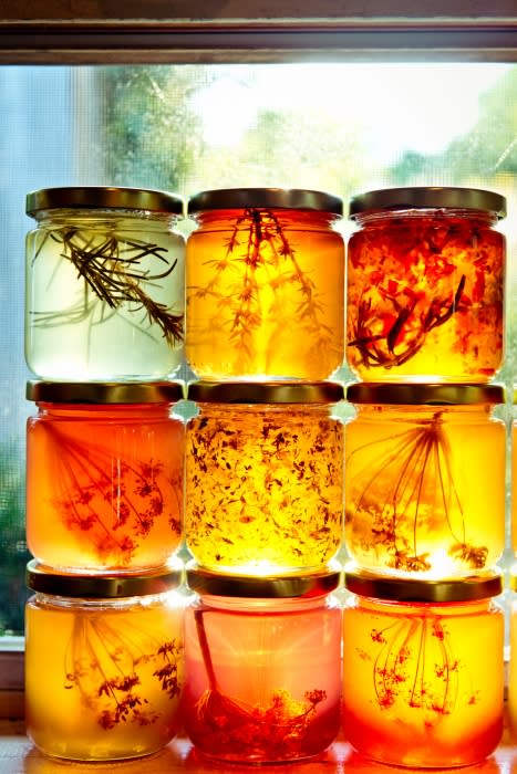 Nine jars of golden honey, each containing different herbs and spices, are stacked in a window with natural light illuminating them.