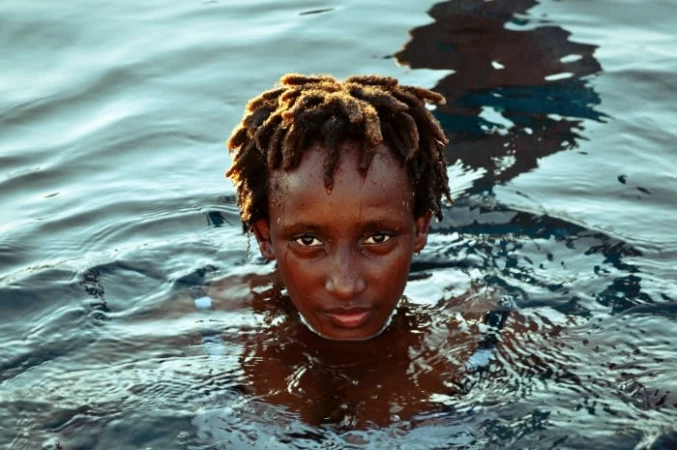 Close-up portrait of a Black person with dreadlocks swimming in dark water, looking directly at the camera