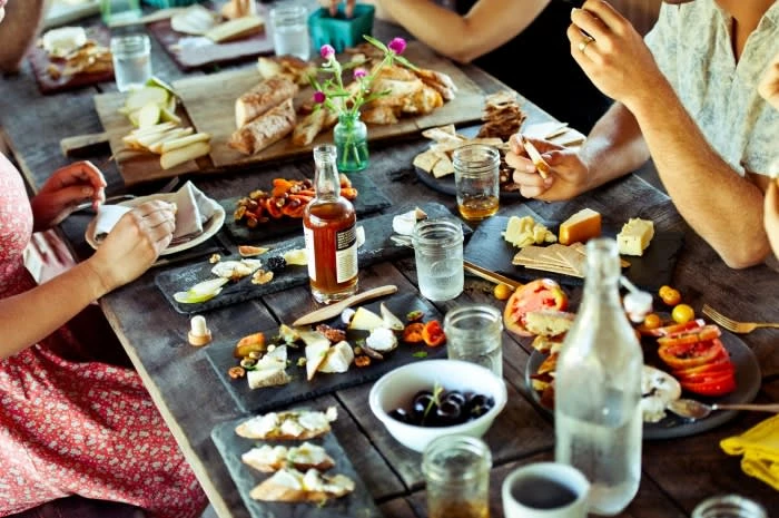 Overhead shot of a wooden table laden with various food items like cheese, bread, and fruit, with several people seated around it.
